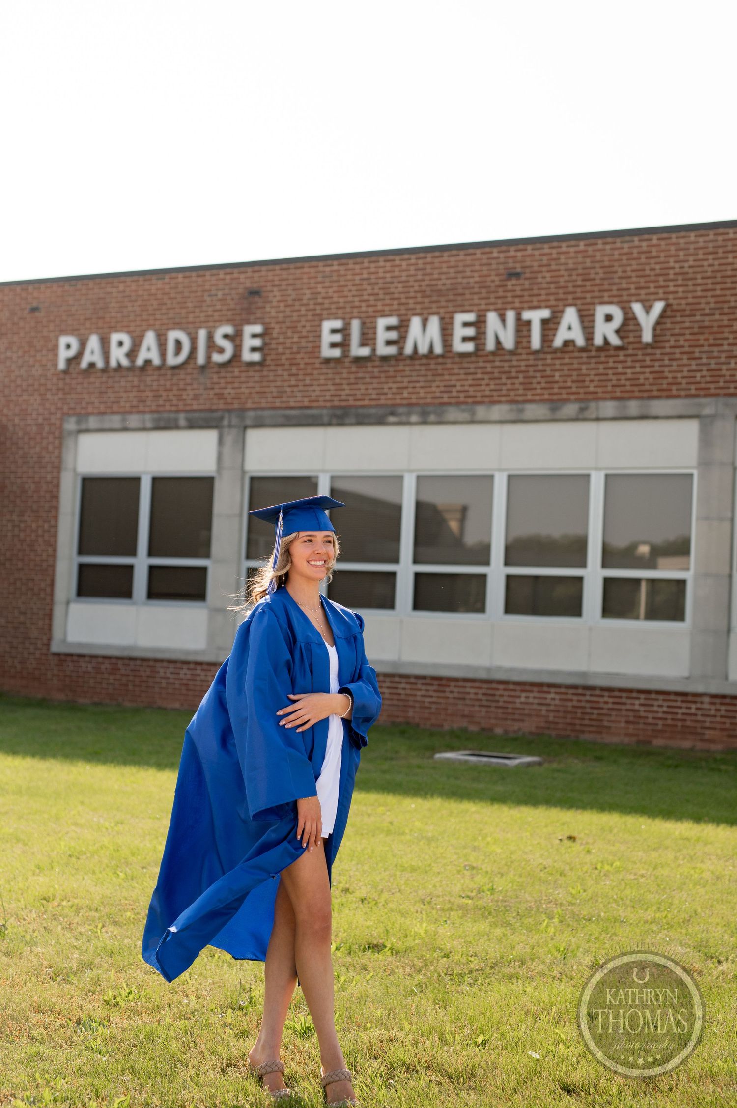 Graduate in blue graduation attire poses confidently outside Paradise Elementary building.