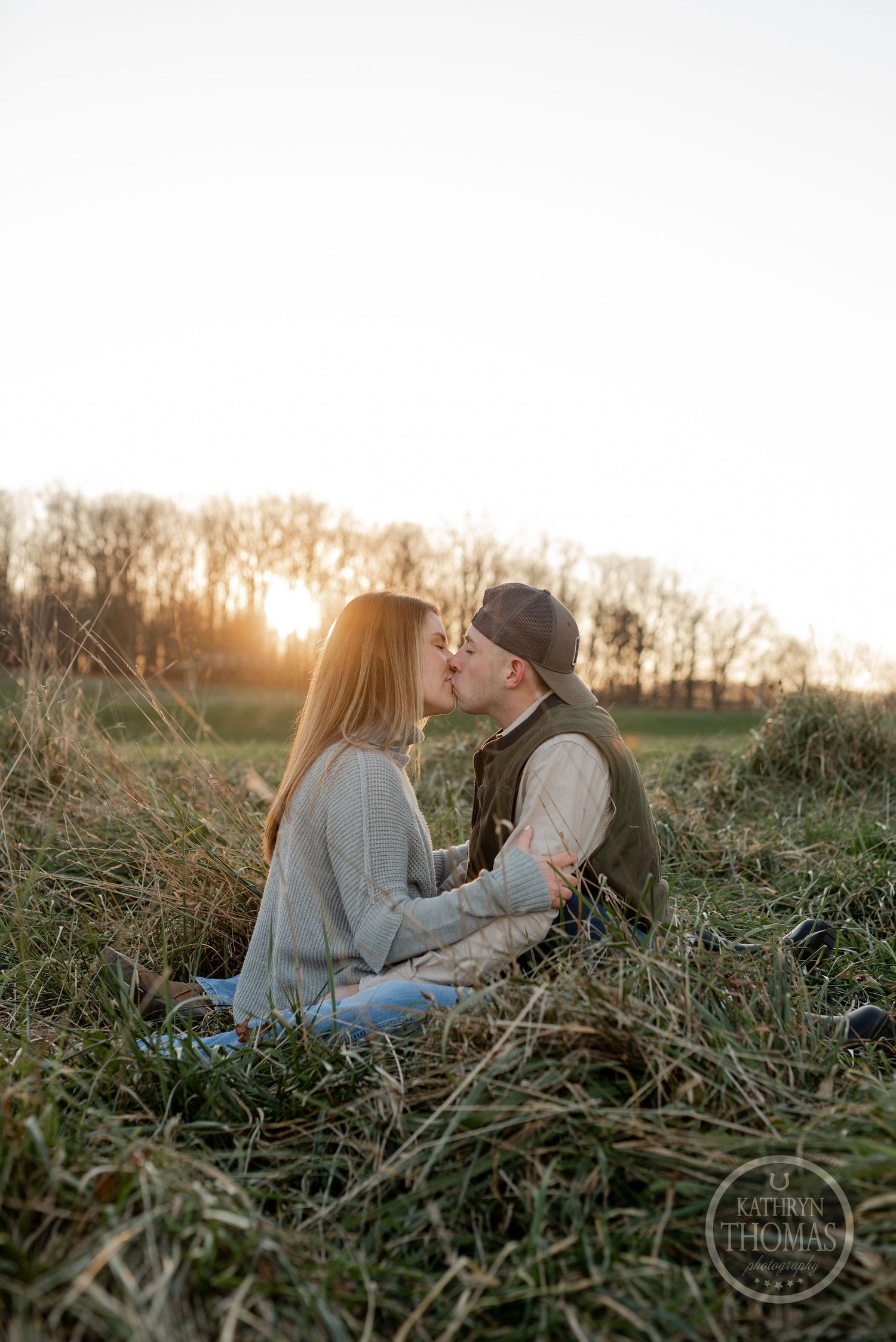 A couple shares a romantic kiss while sitting in tall grass during golden hour.