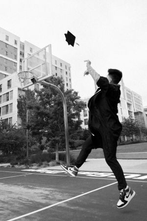 Graduate throws cap in black and white photo on basketball court.