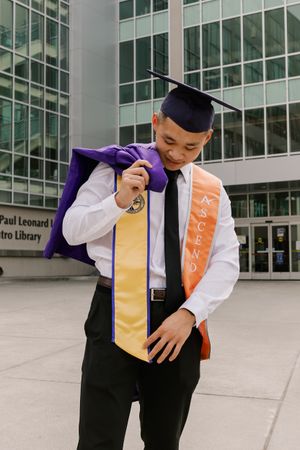 Graduate in academic regalia poses confidently outside a modern glass building.