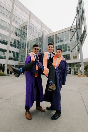 Three graduates in academic regalia pose playfully outside modern campus building.