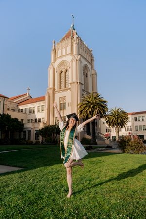 Graduate in white dress and sash leaps joyfully on lawn in front of historic church tower.