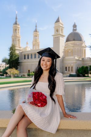 Graduate in white dress holds red flowers while posing by fountain with cathedral building backdrop.