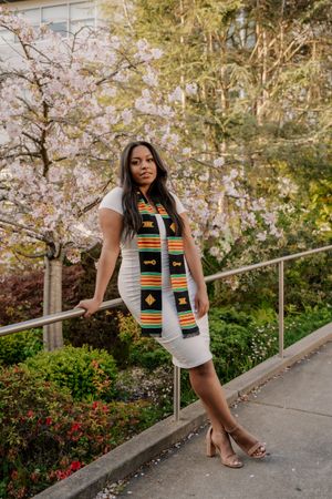 Graduate in white dress and colorful stole poses against blooming cherry blossom tree background.