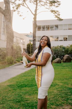 Graduate in white dress with graduation stole celebrates outdoors during golden hour.
