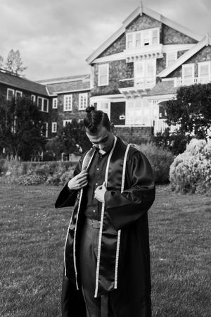 Graduate in black academic regalia poses in front of historic stone building in black and white photo.