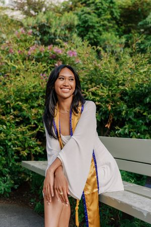 Graduate in white dress and gold sash sits on bench amid flowering garden setting.