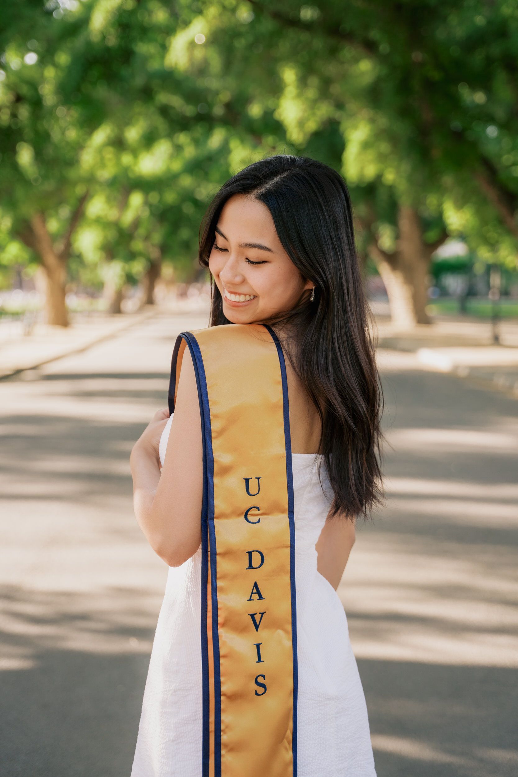 UC Davis graduate displays gold and blue graduation sash in outdoor campus setting.