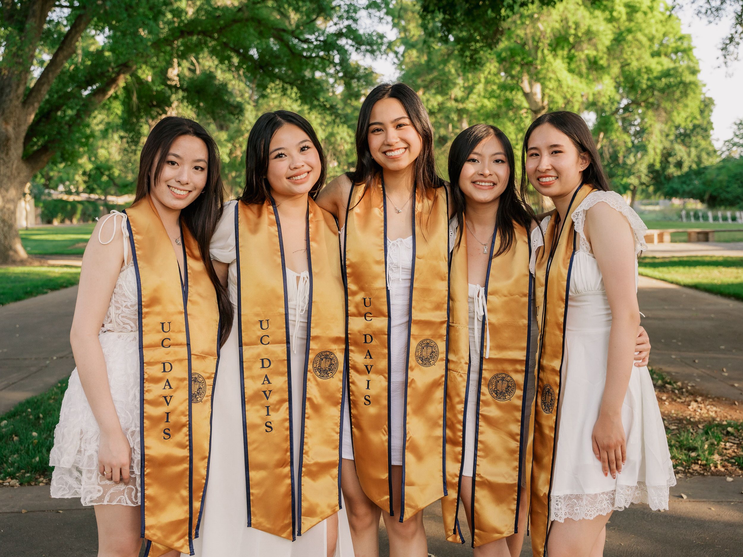 Graduates in white dresses wearing gold sashes pose together on green campus lawn.