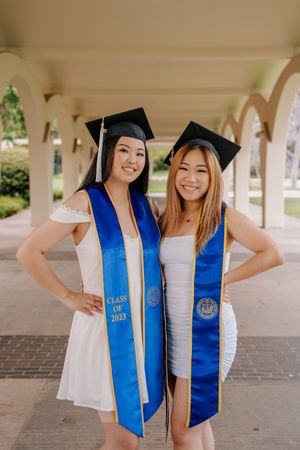 Two graduates in white dresses with blue graduation stoles pose under campus archway.