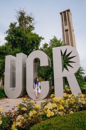 Graduate poses with large UCR letters and campus bell tower in background.