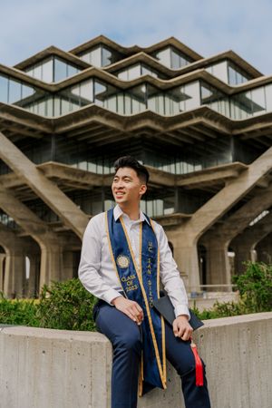 Graduate in formal attire poses in front of distinctive brutalist campus architecture.