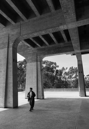 Black and white photo of graduate walking under concrete university building architecture.