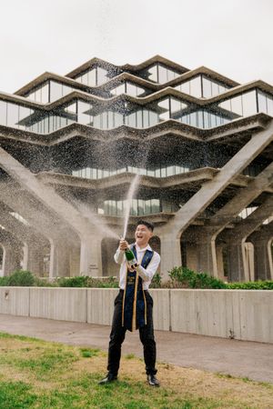 Graduate in cap and gown stands beneath unique zigzag concrete architecture while spraying water.