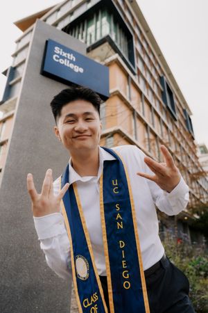 Graduate in white shirt and blue sash poses in front of Sixth College building.