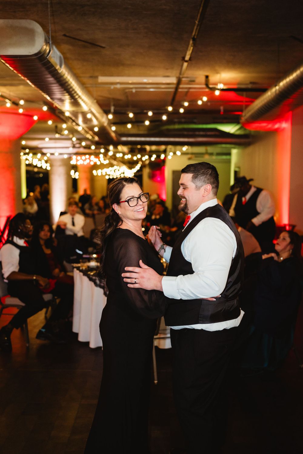 A couple dancing together at an indoor reception venue with red accent lighting and exposed ceiling.
