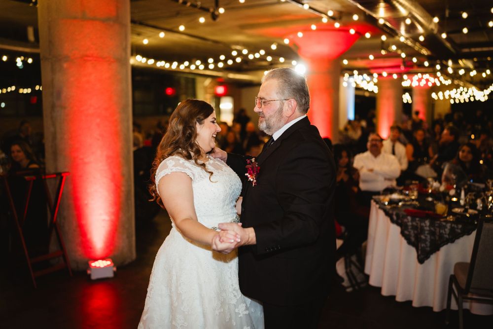 A couple shares their first dance at a wedding reception under string lights with red-lit columns in an industrial venue.