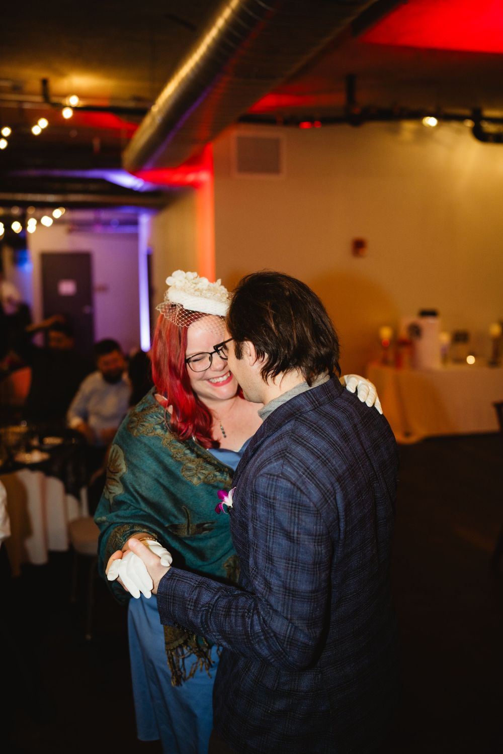 Two people share an intimate dance moment at a dimly lit indoor reception with colorful lighting.