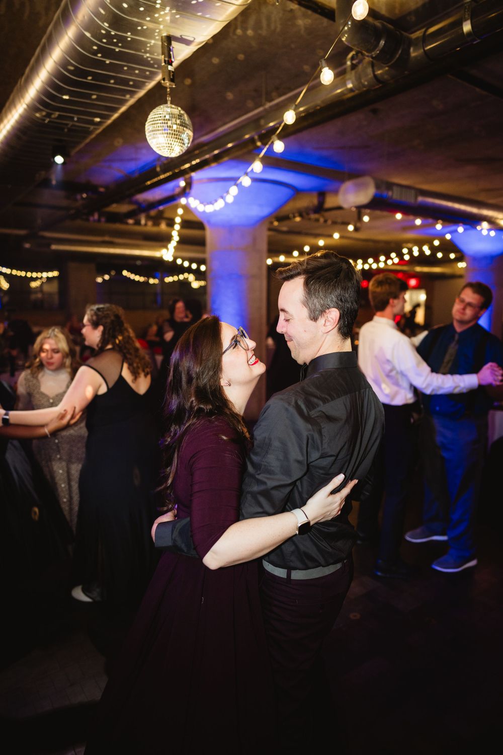 Couples dancing under string lights and disco ball at an indoor celebration with industrial decor.
