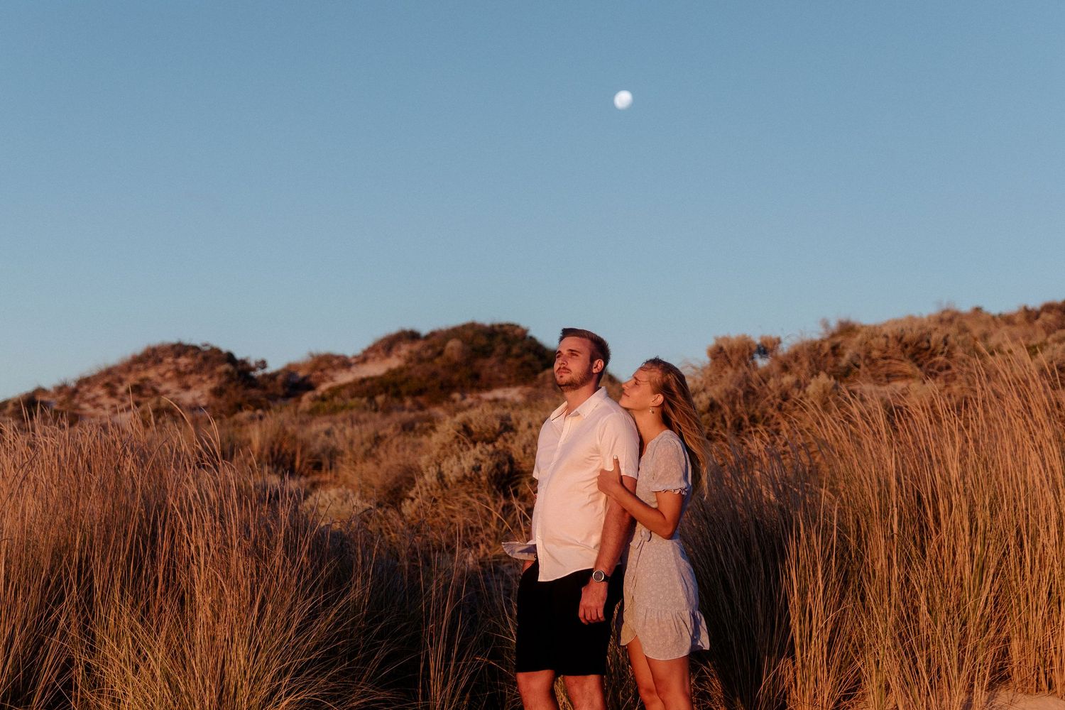 A couple stands together in tall golden grass at sunset with a moon visible in the blue sky.