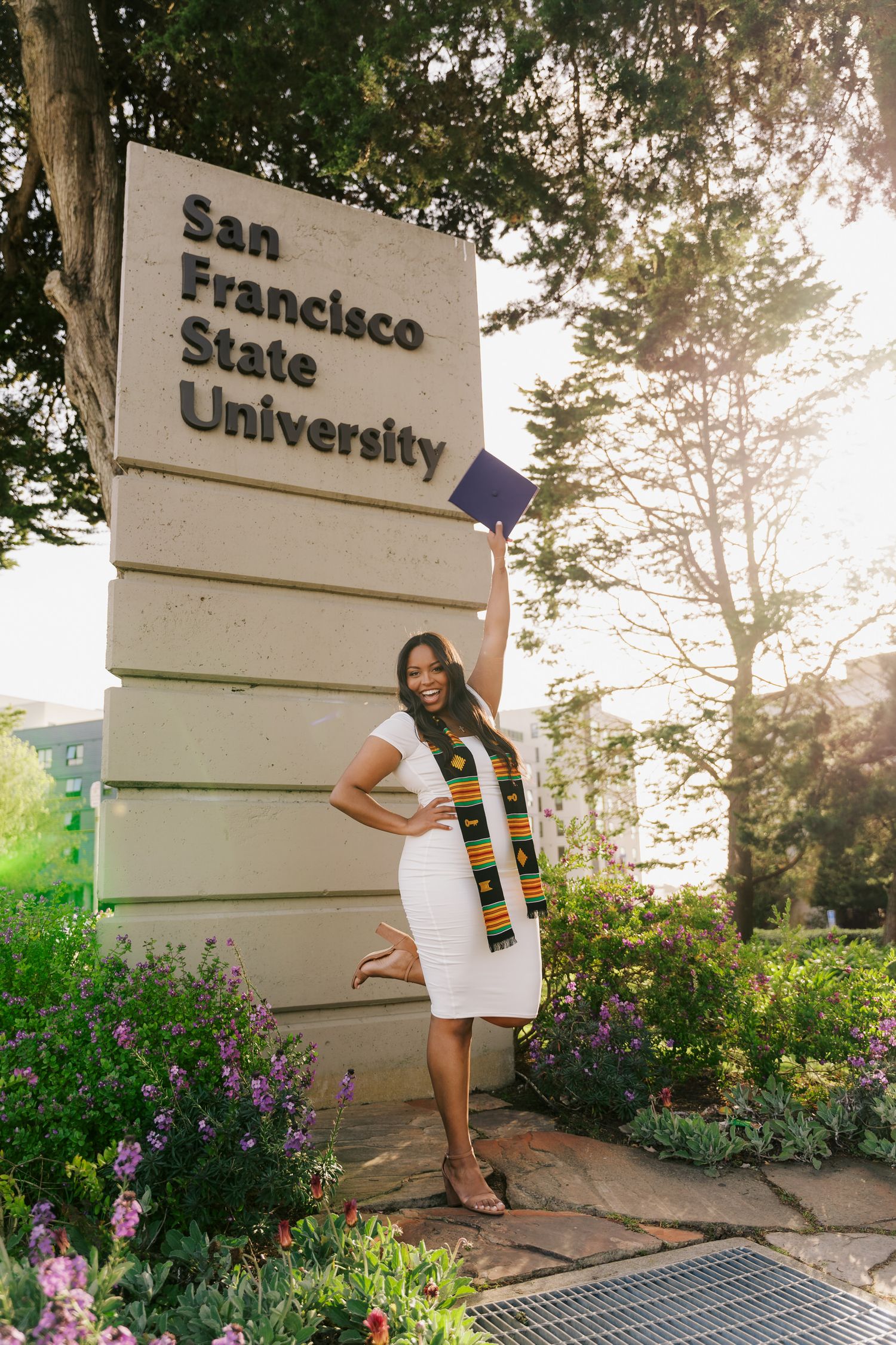 Graduate posing next to San Francisco State University entrance sign surrounded by flowers.