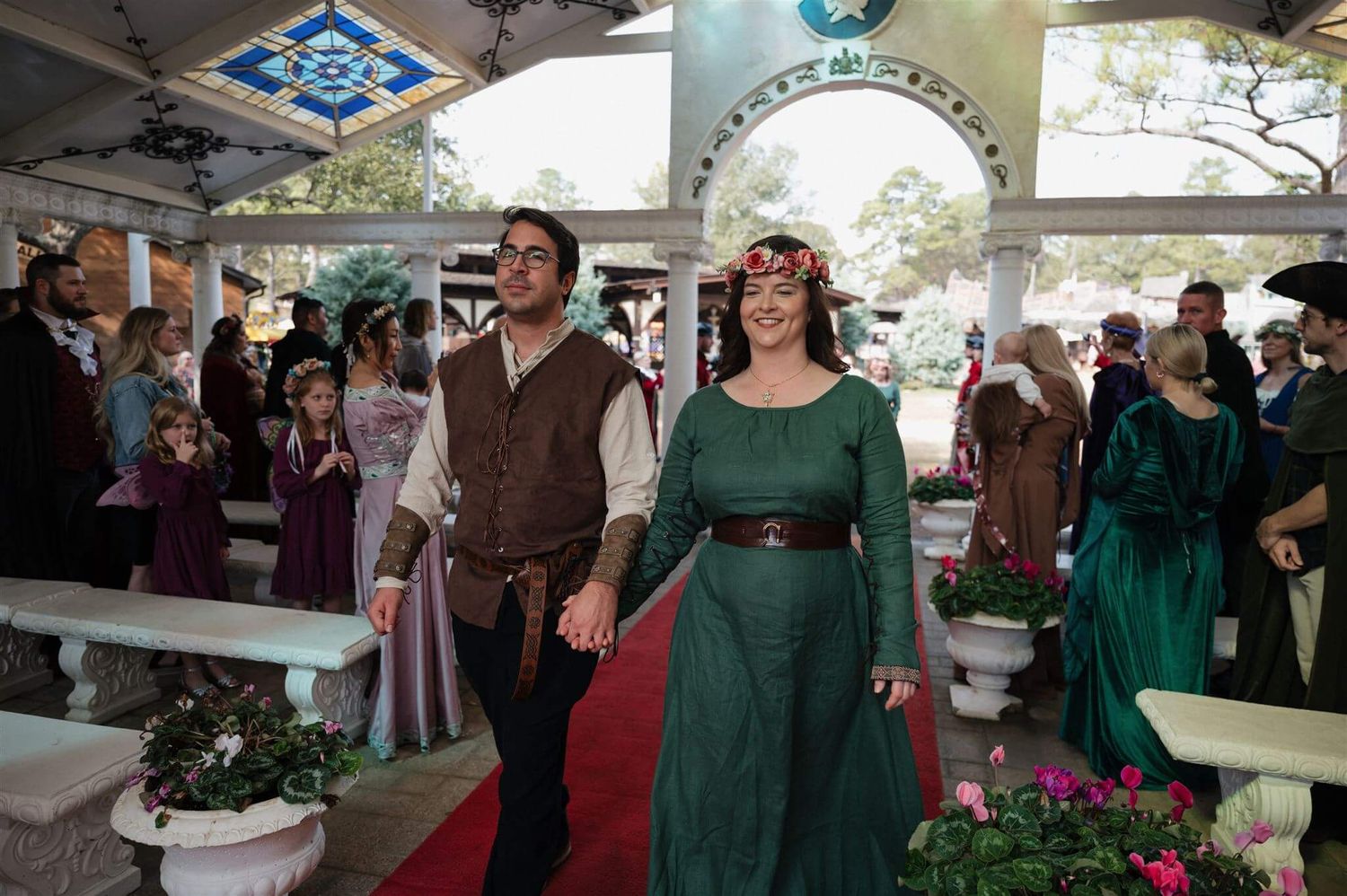 Couples in medieval costumes walking down a red carpet aisle during a renaissance ceremony.