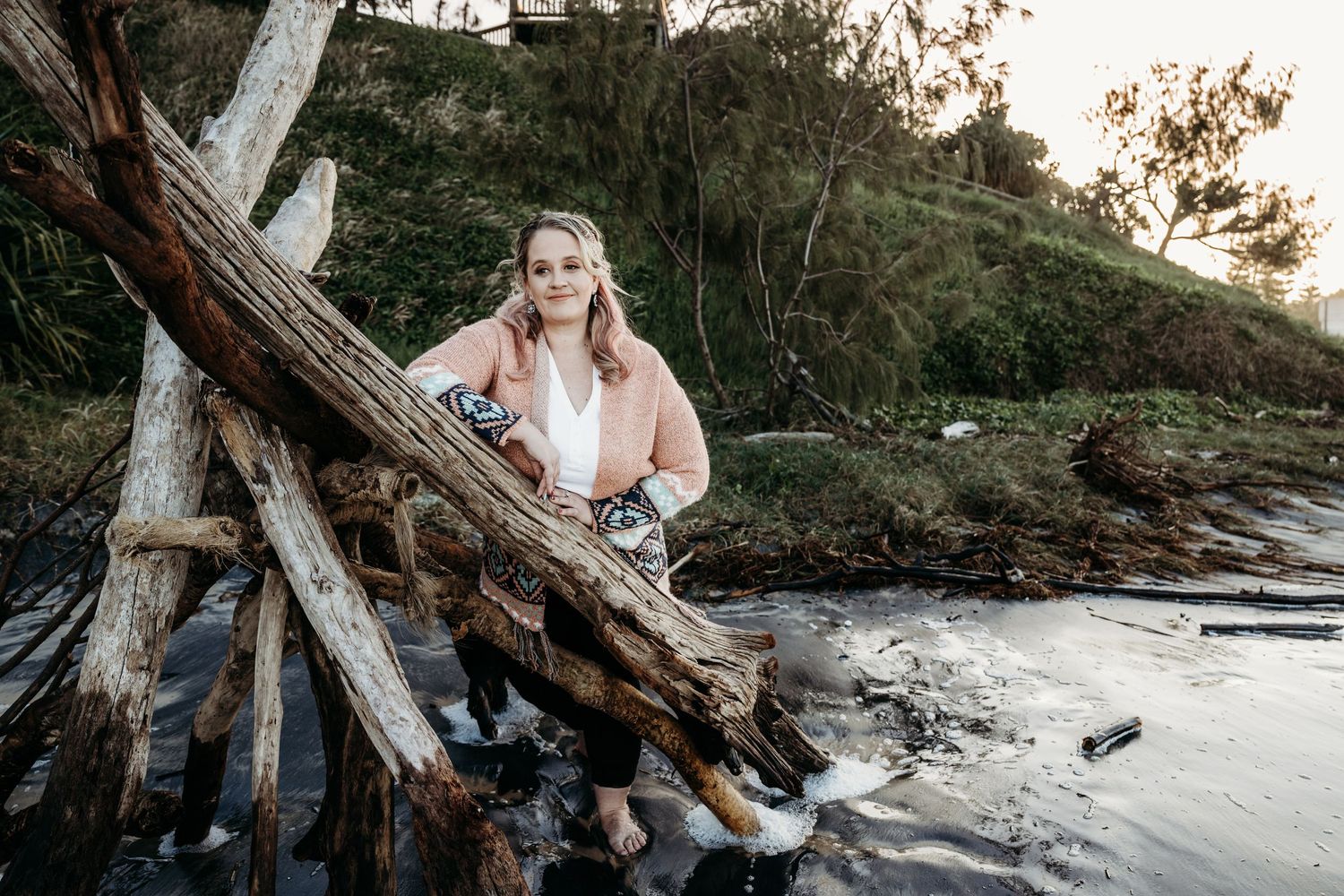 A dreamy coastal portrait with driftwood and rocks along a serene beach at sunset.