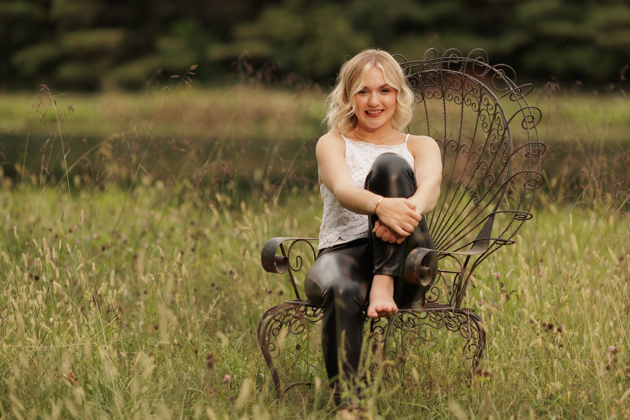 A pretty, young lady sits in a wrought iron chair in front of a wooded pond during summer.