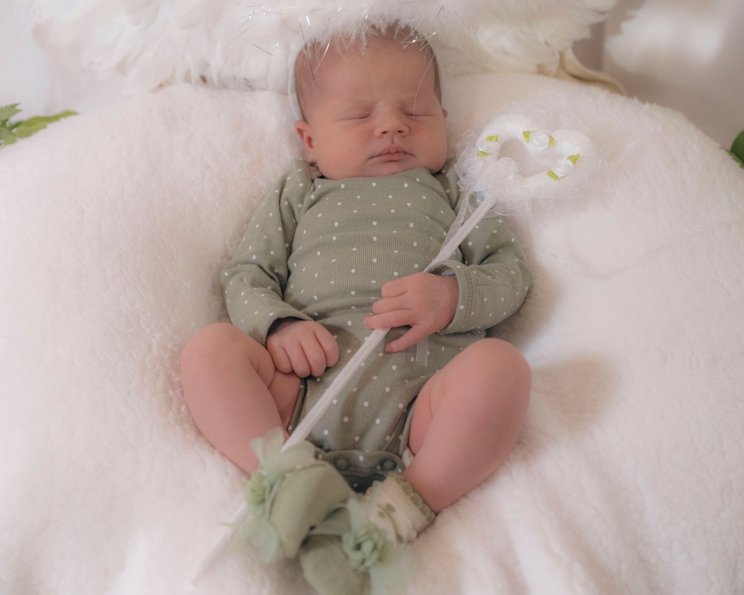 Close-up of sleeping newborn in green polka dot onesie holding a fairy wand on white fur.