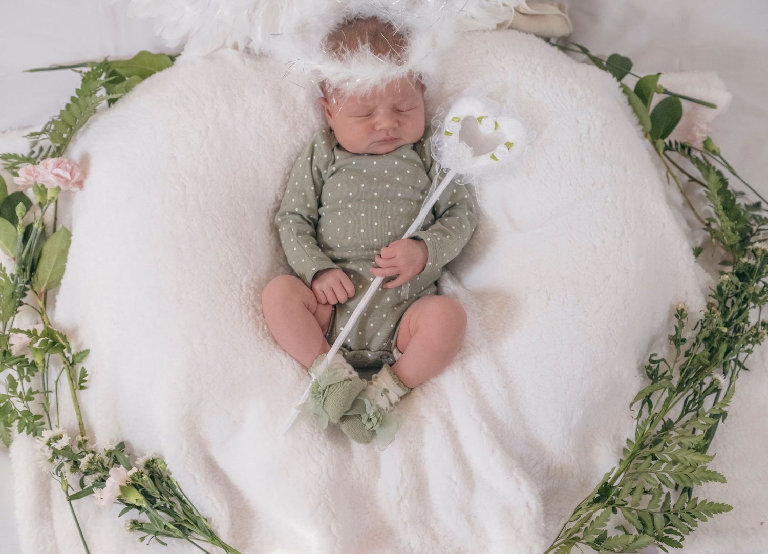 Serene newborn angel in green onesie with halo headband holding a wand in a flower wreath.