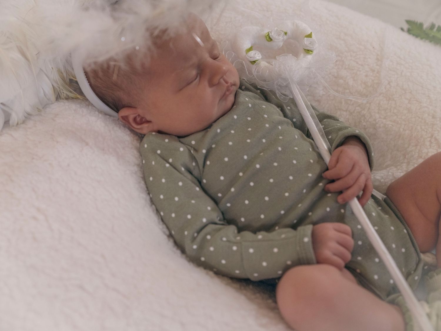 Close-up of a sleeping newborn in green polka dot onesie holding a white wand with floral headband.