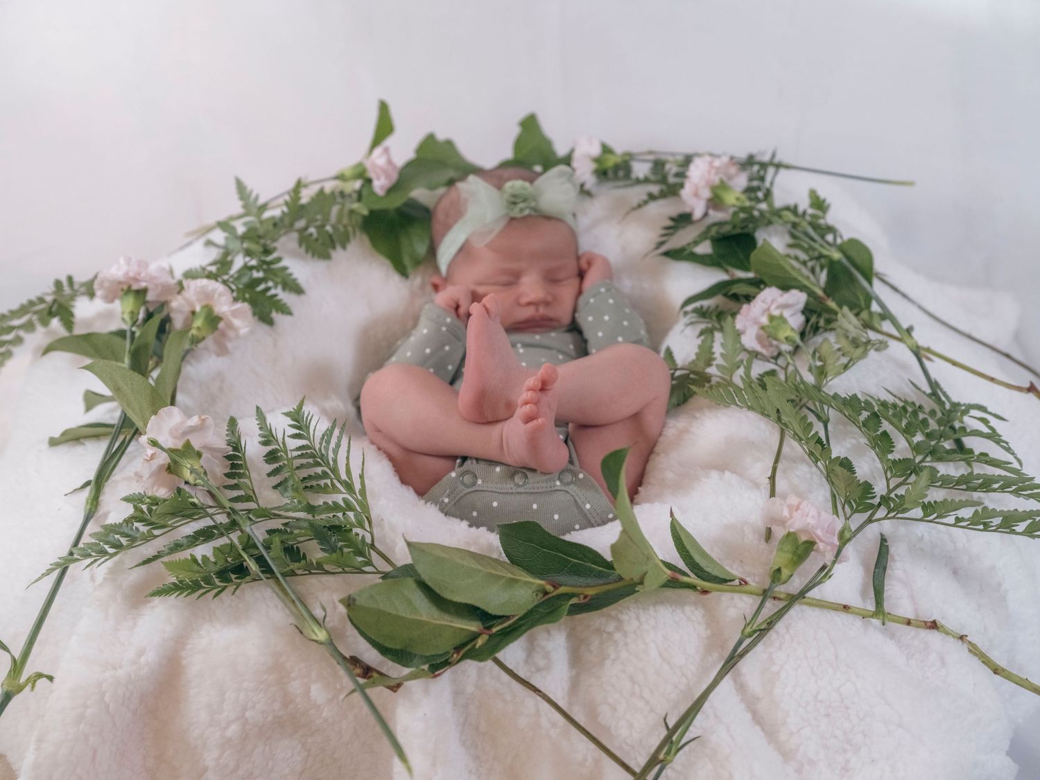 Newborn baby in grey onesie sleeping in a nest of ferns and pink flowers on a white blanket.
