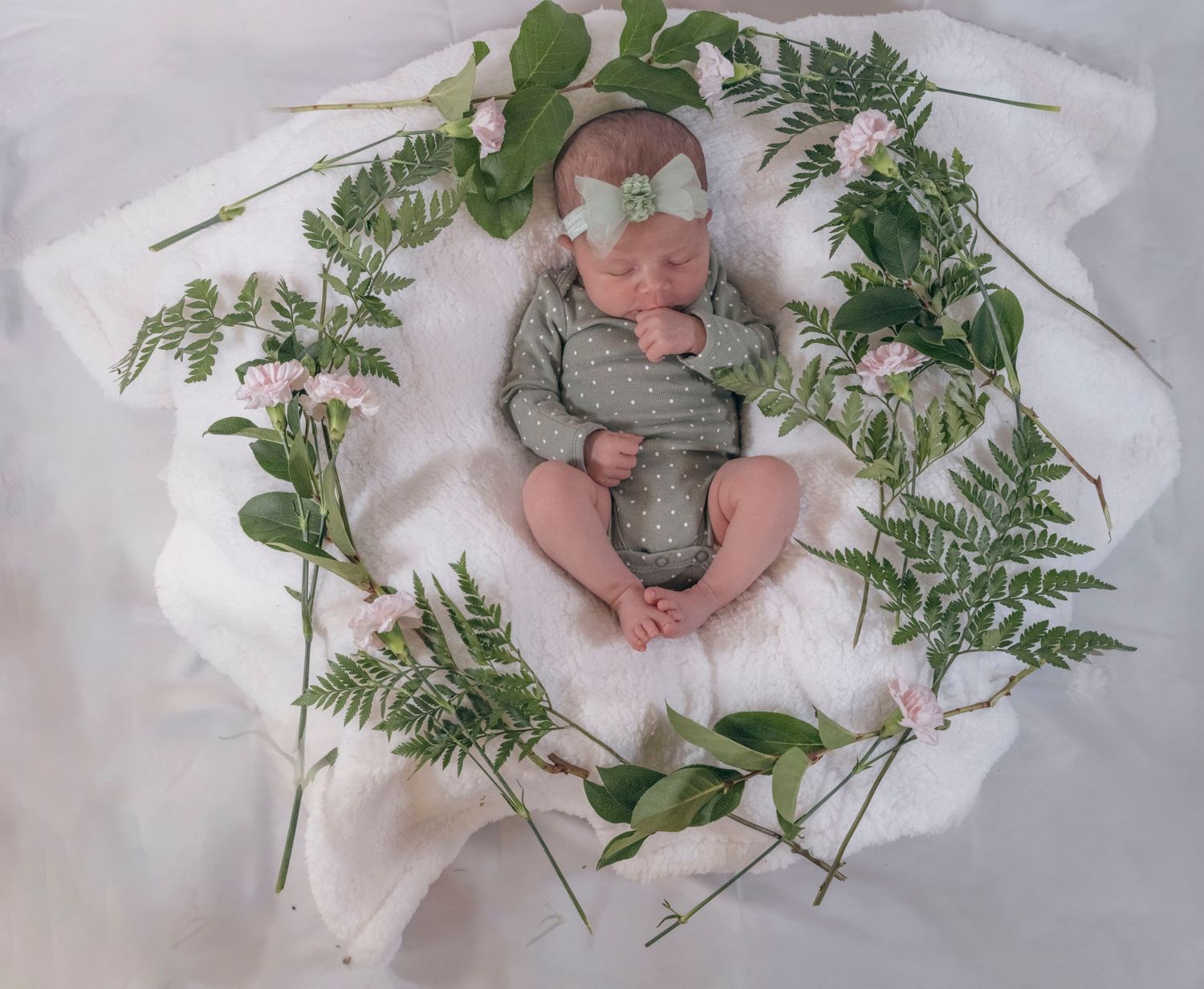 Sleeping newborn in green onesie with bow headband holding a cross, surrounded by ferns and pink flowers.