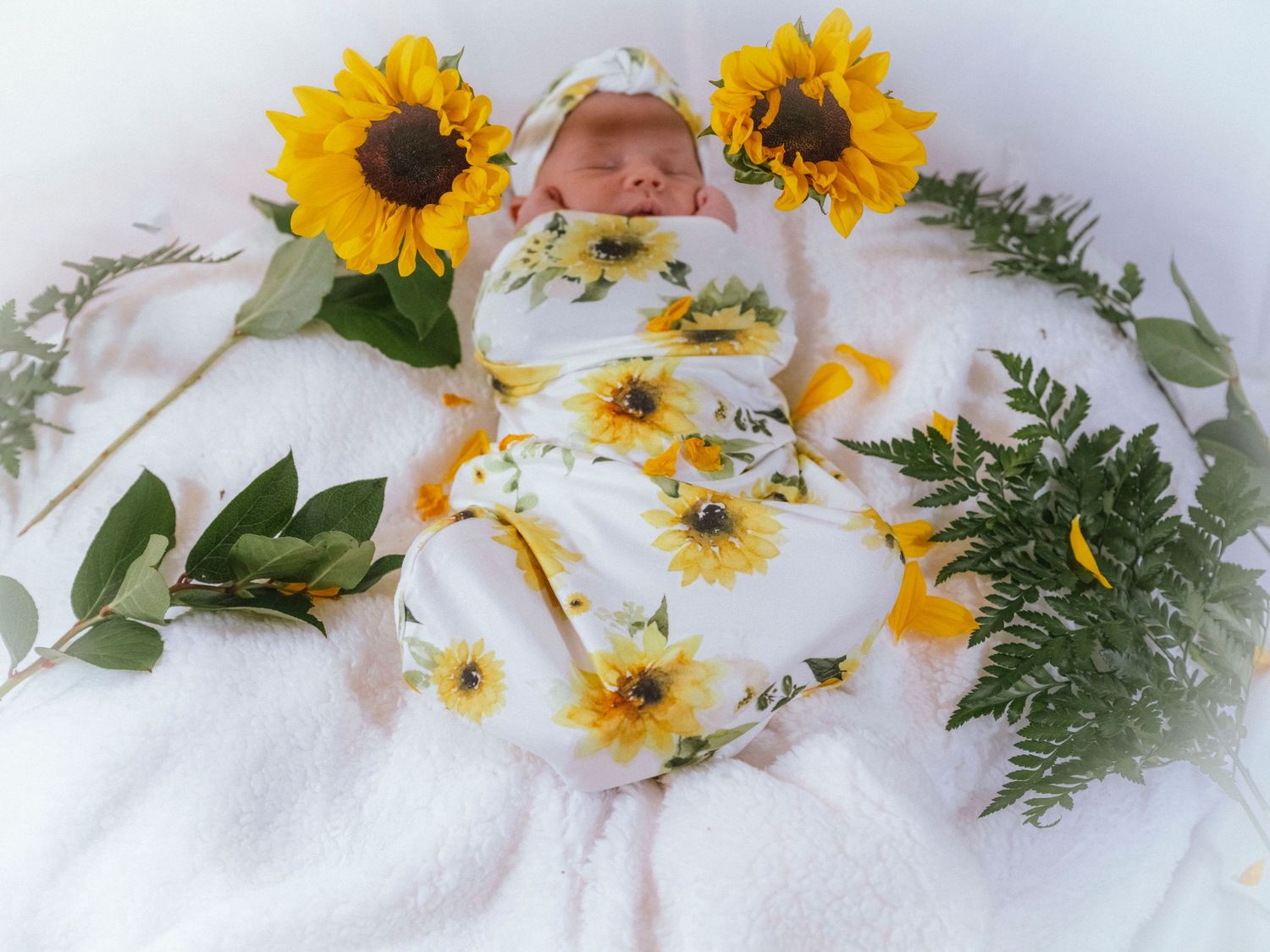 Swaddled newborn baby in sunflower print wrap surrounded by bright yellow sunflowers on white blanket.