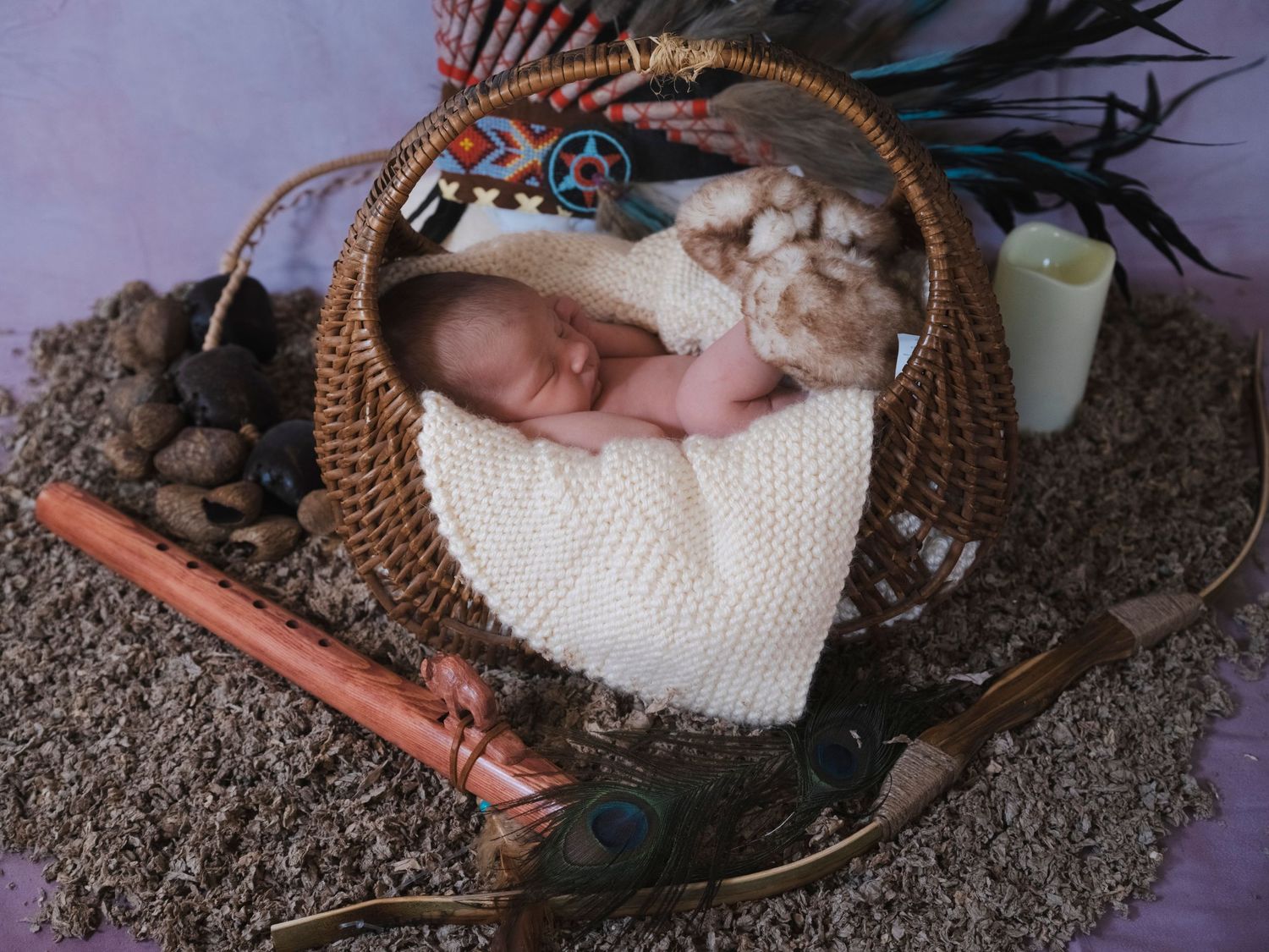 Peaceful newborn baby sleeping in wicker basket surrounded by Native American cultural props and blue feathers.