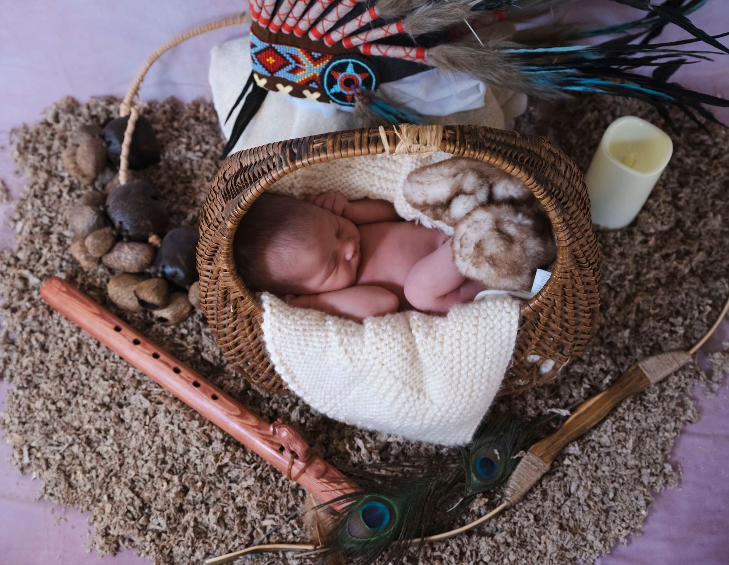Sleeping newborn baby nestled in wicker basket with Native American feathers, flute, and decorative props.
