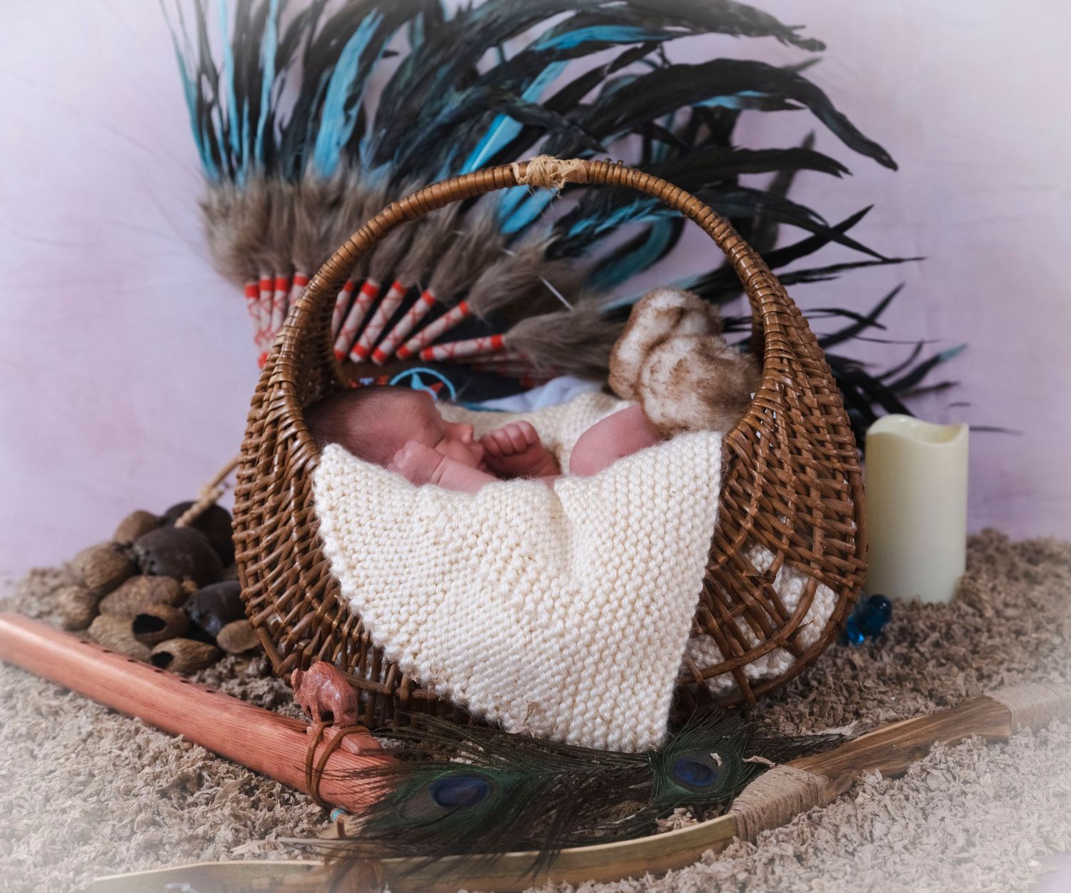 Sleeping newborn baby wrapped in cream knit blanket in wicker basket with blue feathers and candle.