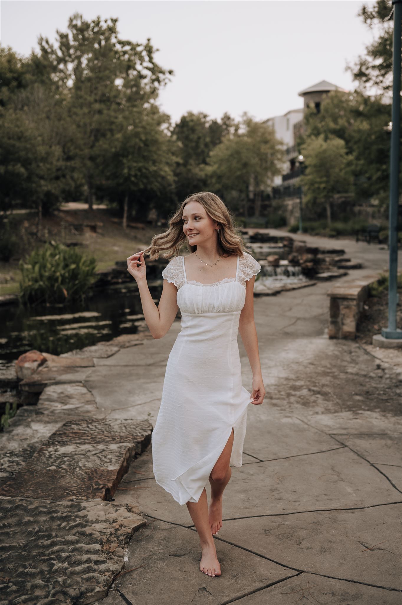 A joyful pose in a white dress along a stone pathway near a decorative water feature.