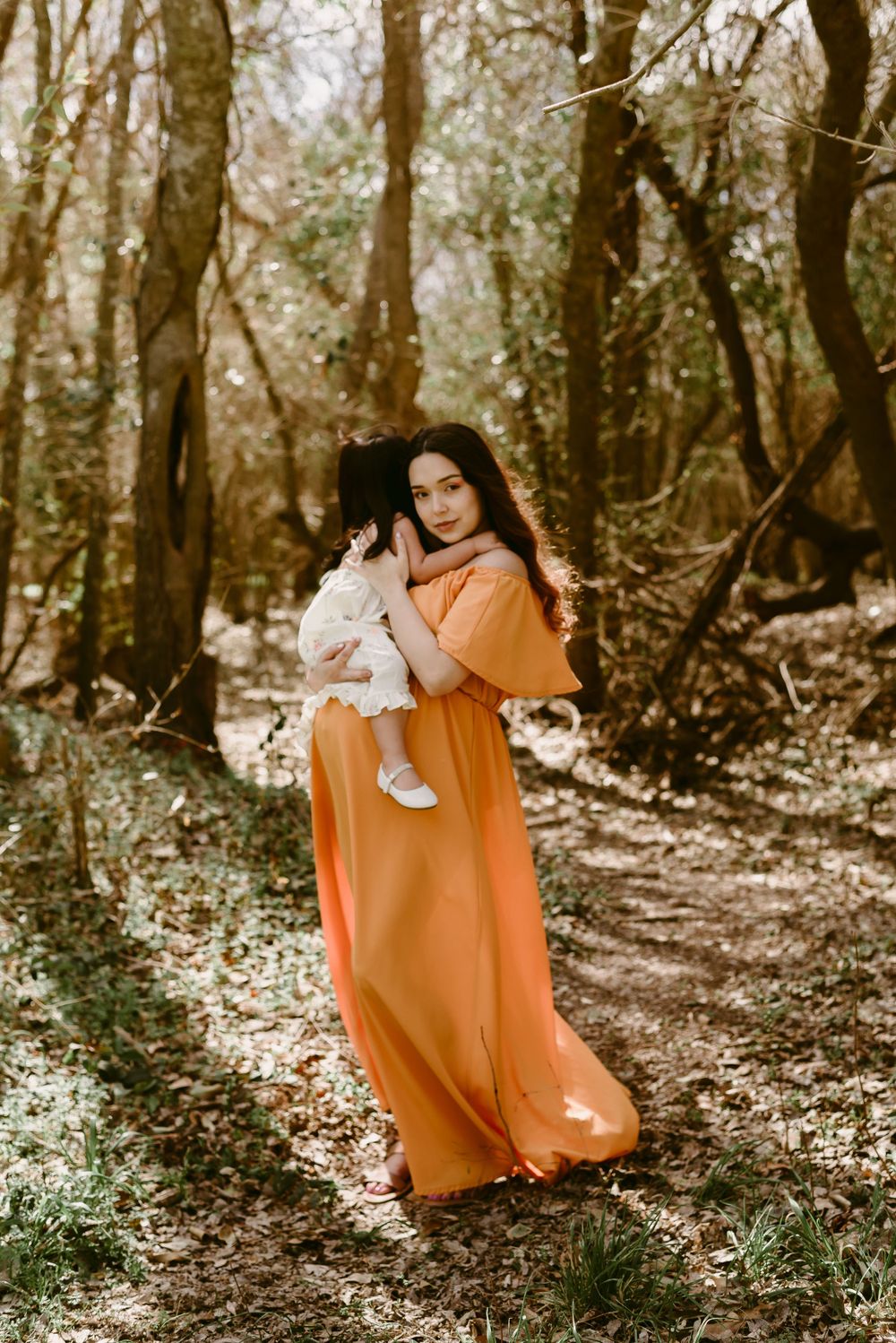 Woman in flowing orange dress holds small child against wooded natural backdrop.