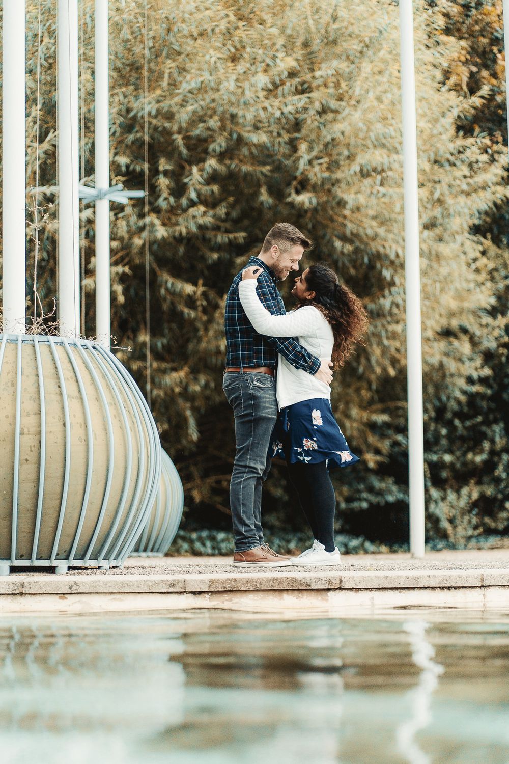 A romantic couple embraces by a decorative gazebo near a reflective pool surrounded by autumn foliage.