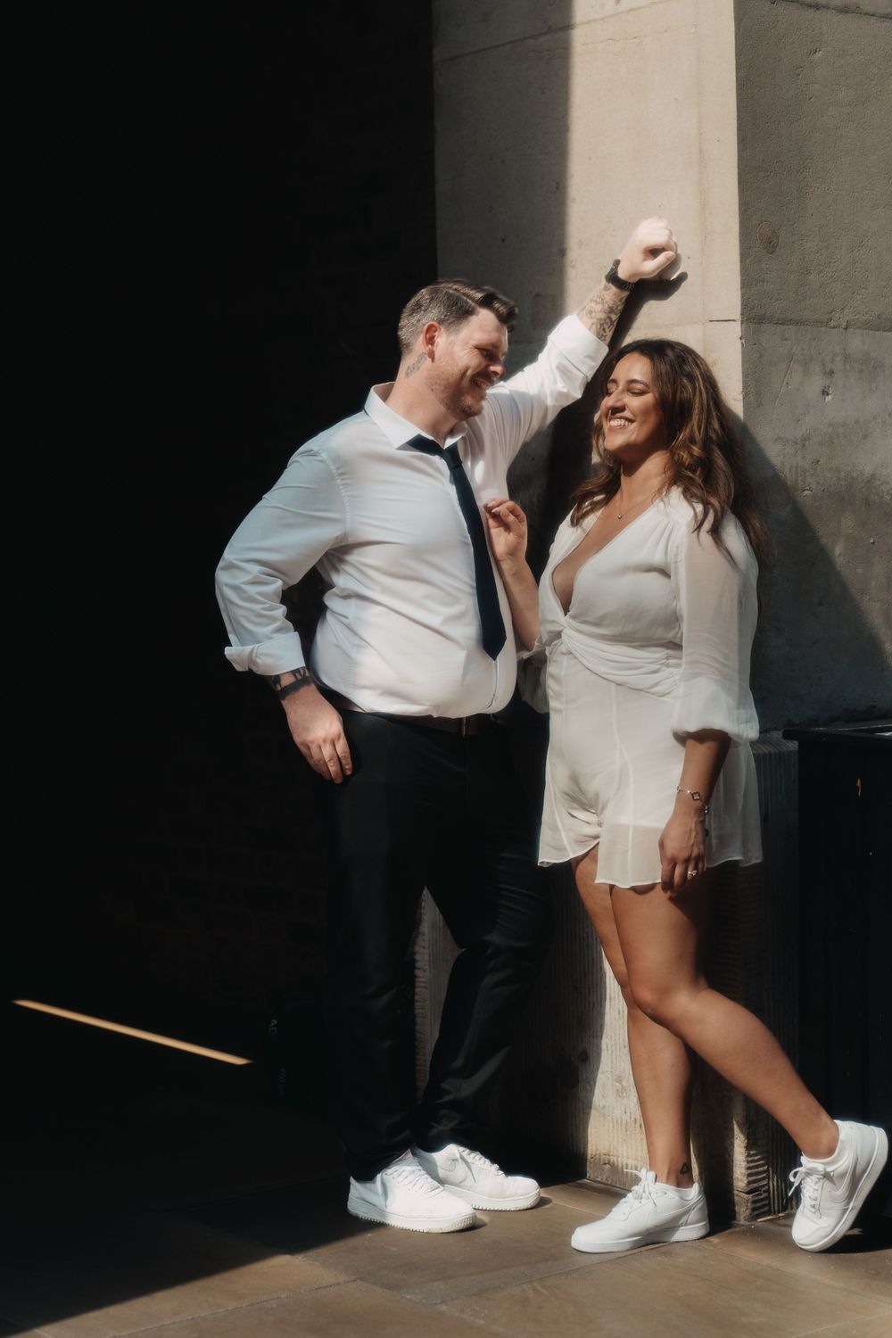 Couple in coordinated white outfits leaning against a pillar in an urban setting.