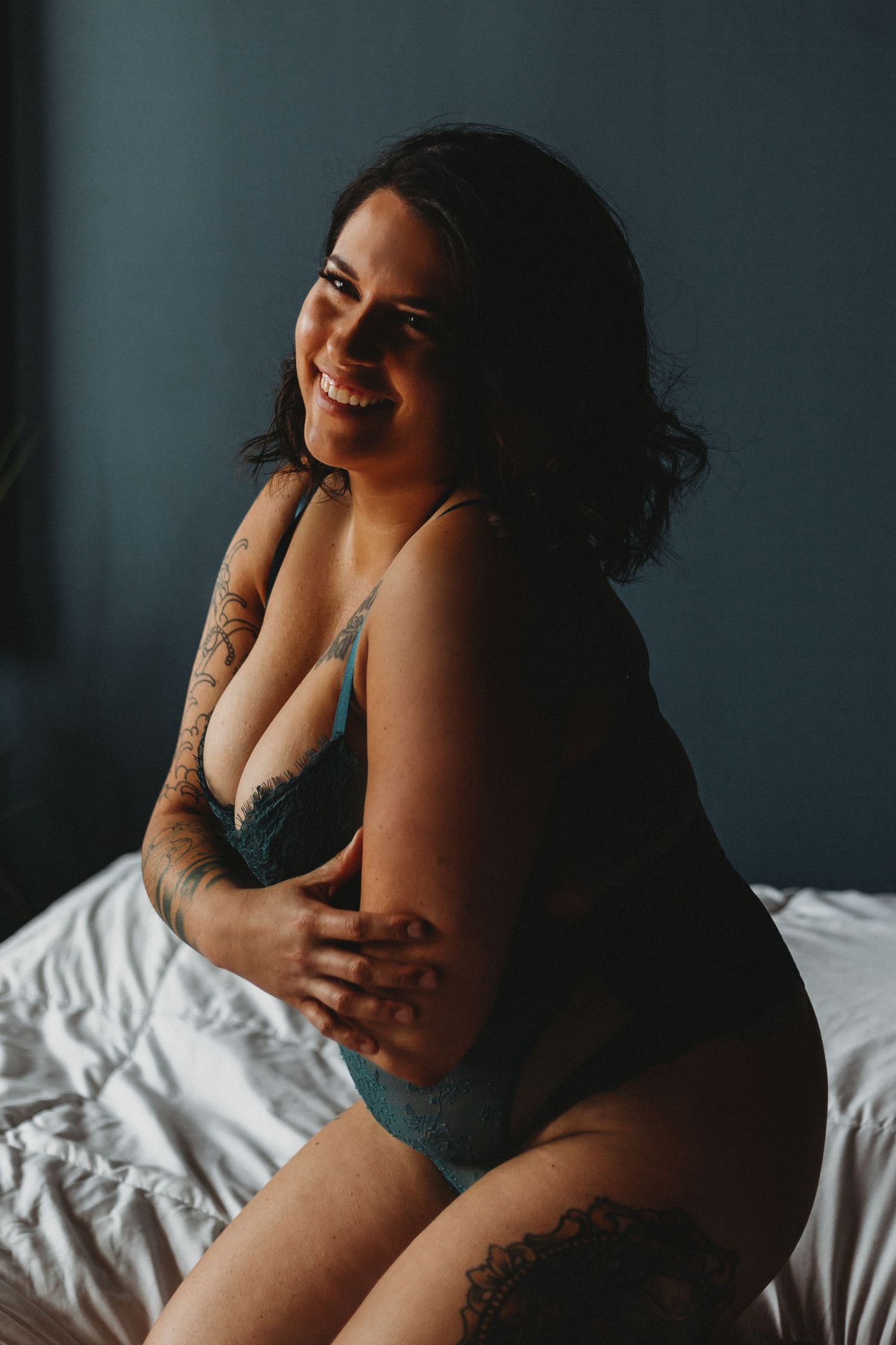 A person in teal lingerie sits on white bedding while smiling in a moody boudoir photoshoot.