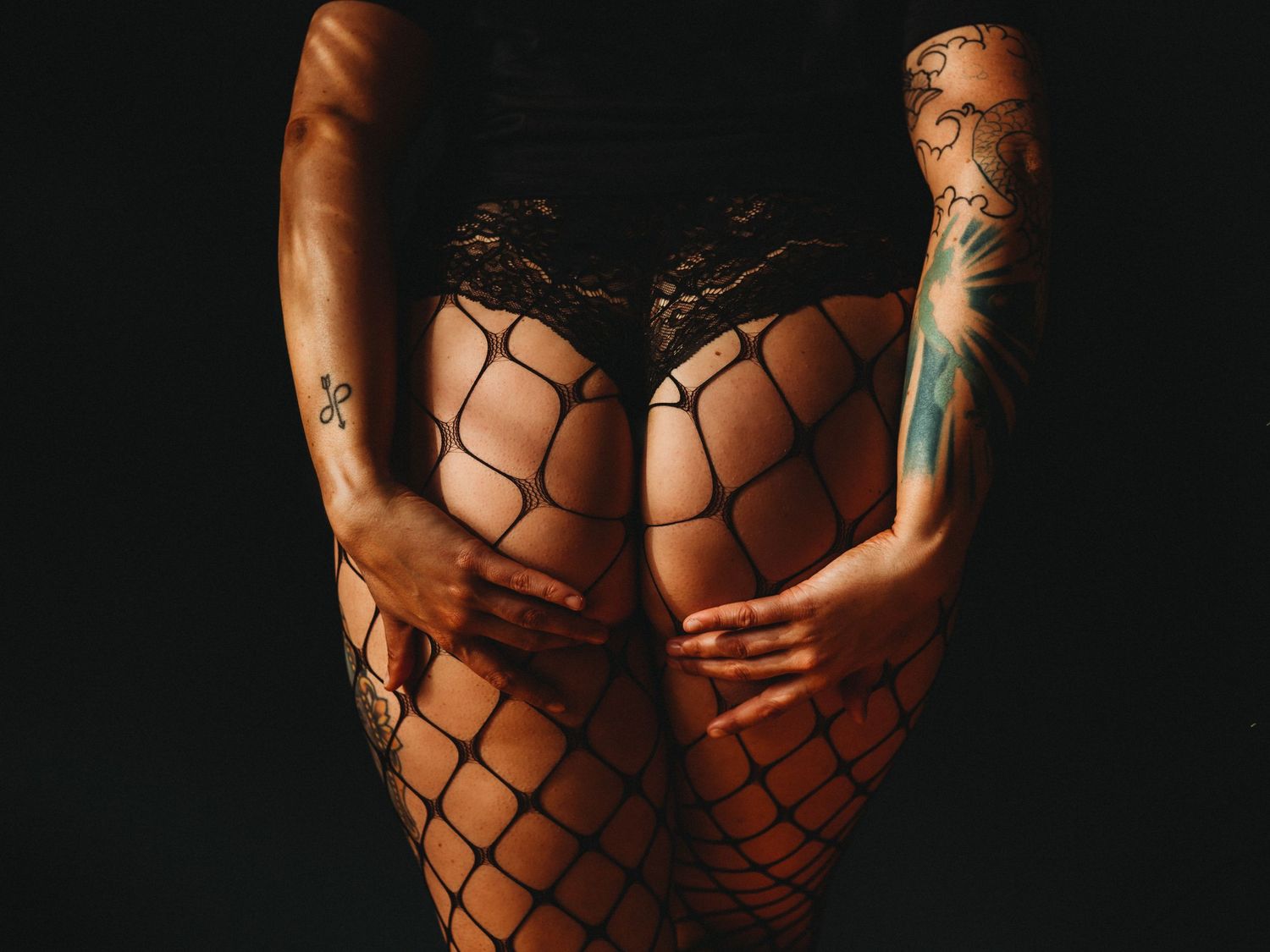 Artistic photo showing tattooed arms and fishnet stockings against dark background.