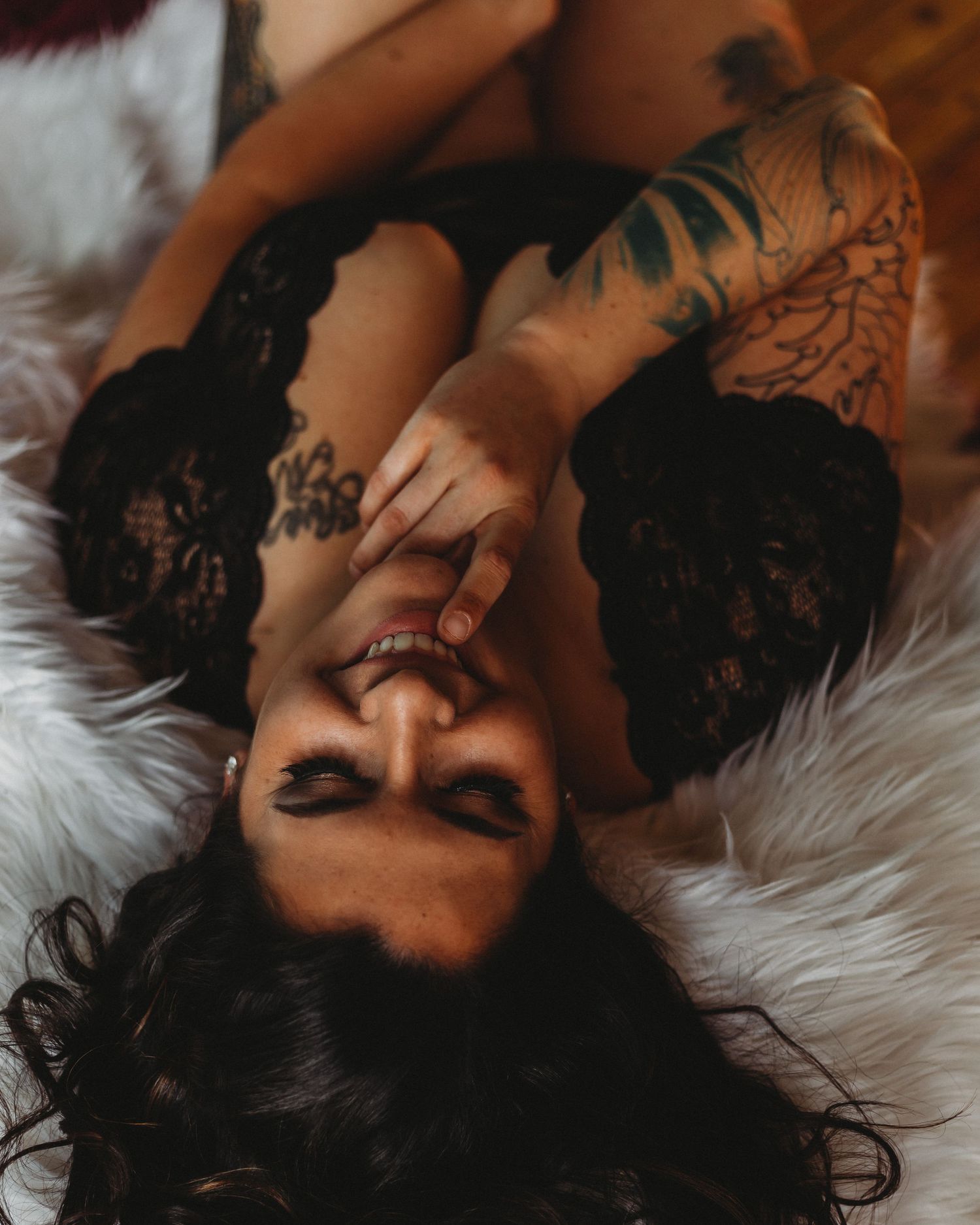 Portrait in soft lighting shows figure laying on white fur with black lace lingerie and artistic tattoos.