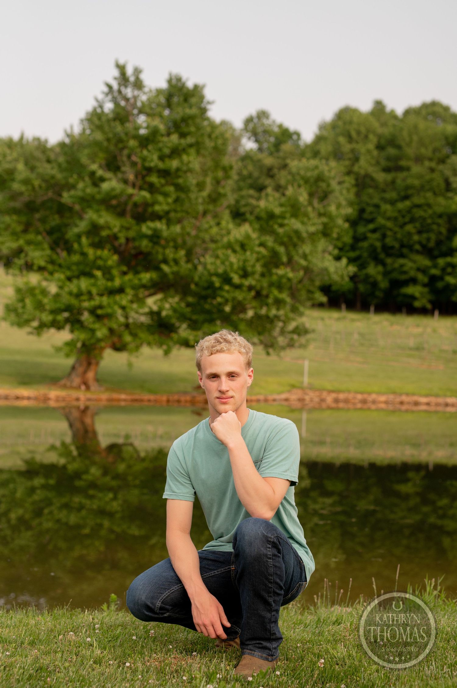 Person crouches thoughtfully by pond with tree reflection in rural setting.