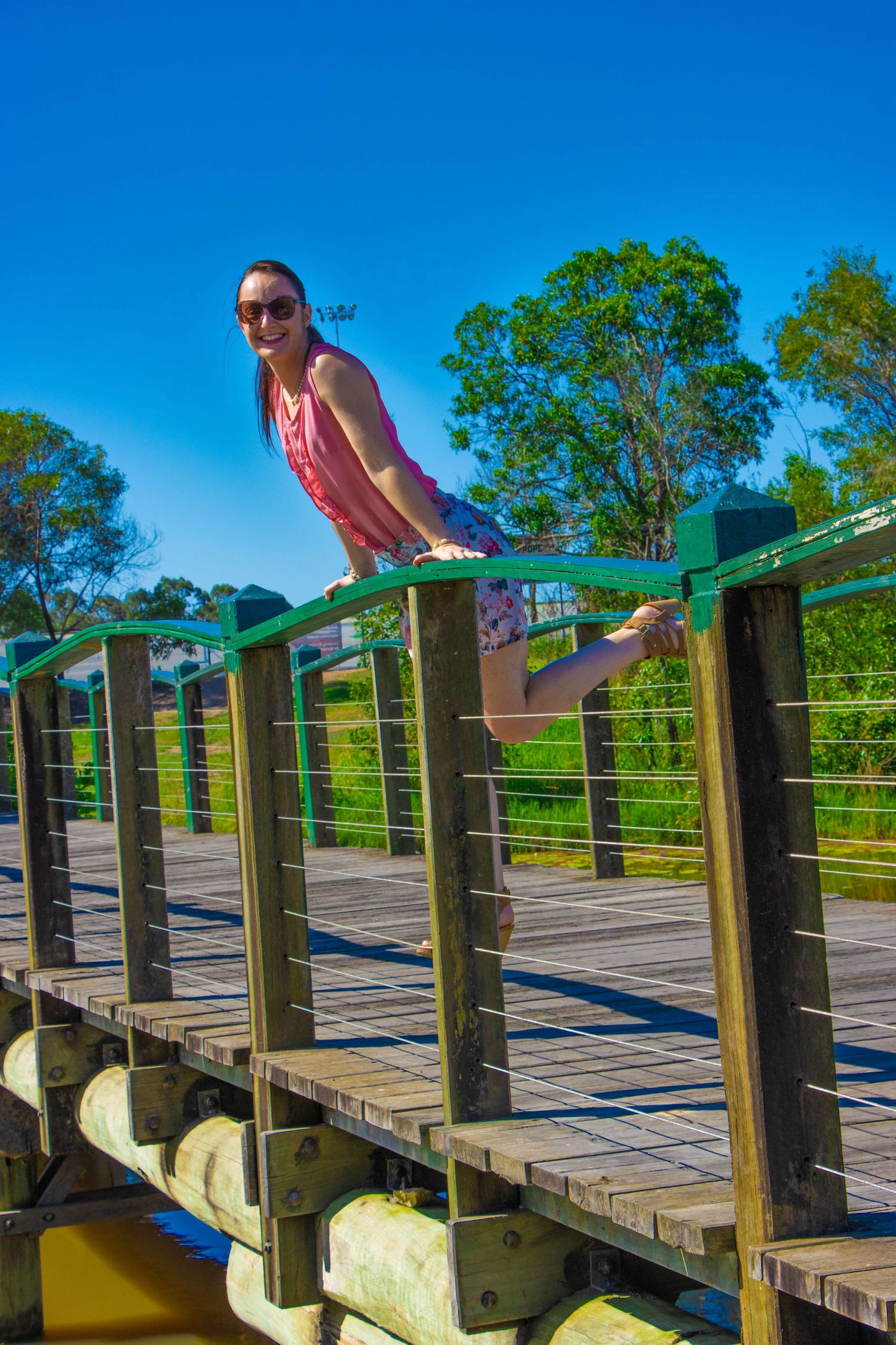 Playful pose on wooden boardwalk bridge against bright blue sky and green trees.