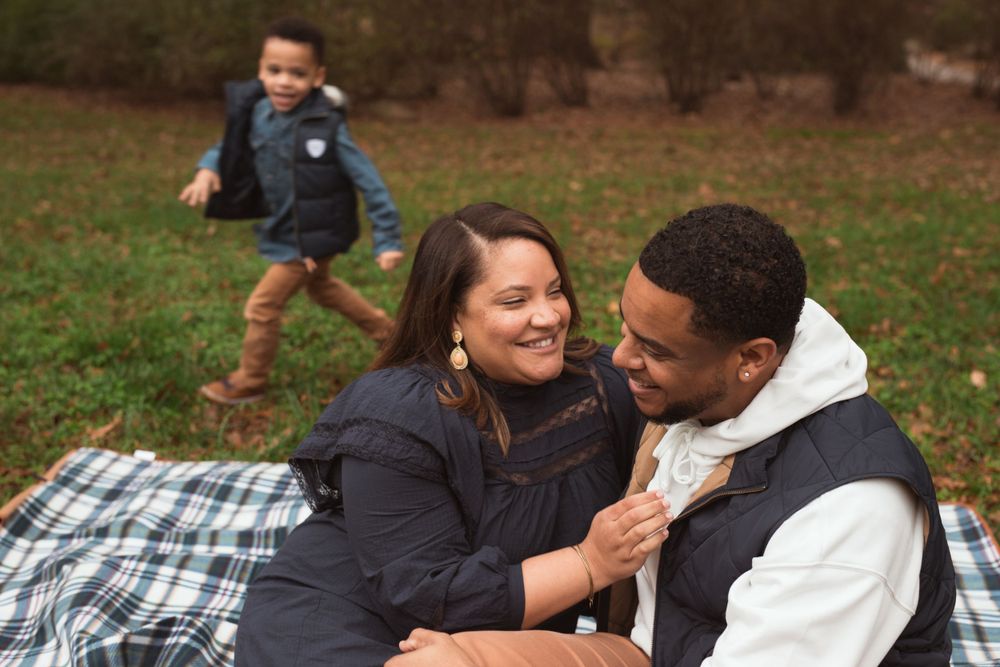 black couple laughing while sitting on a picnic blanket with child playing behind them