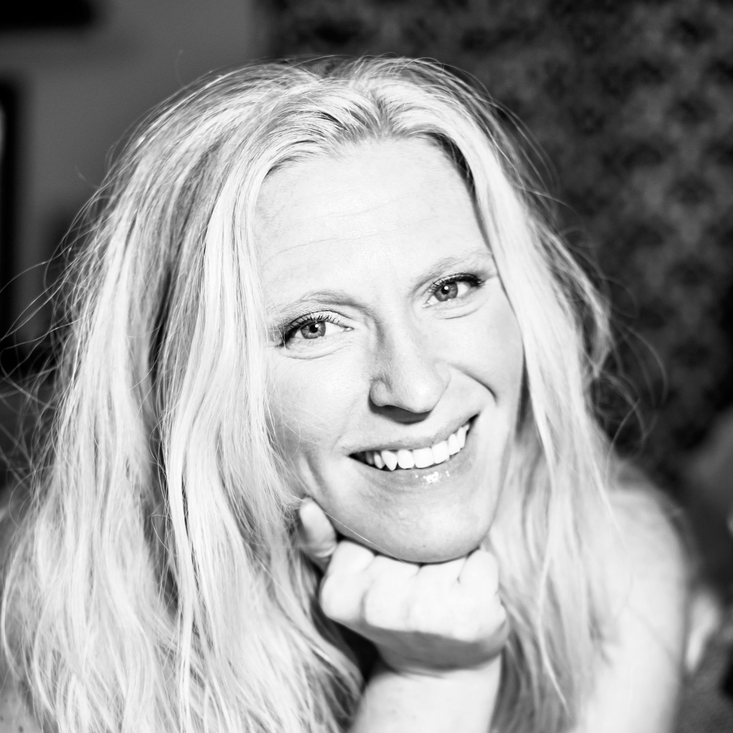 Black and white portrait photograph of a smiling person with long blonde hair resting their chin on their hand.