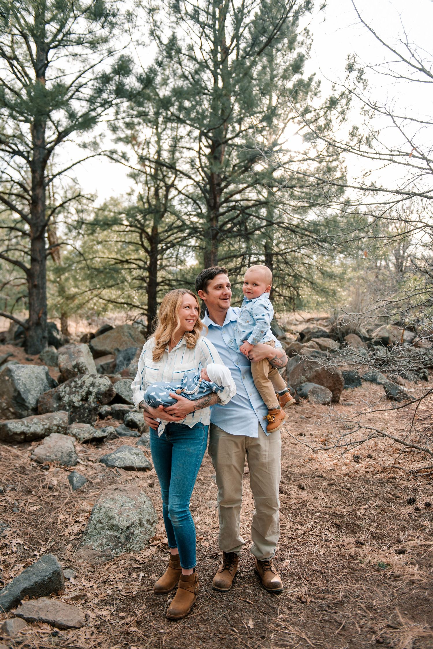 Family walks together among pine trees and rocky terrain during outdoor photo session.