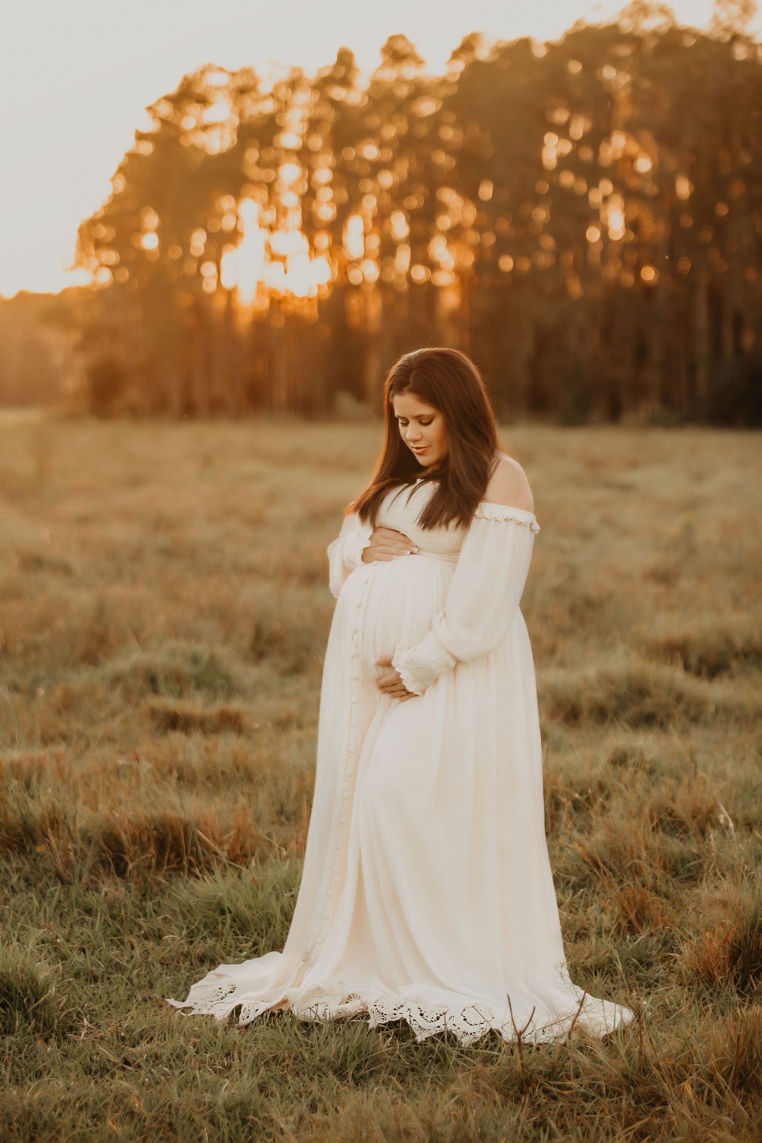 Ethereal maternity portrait in flowing white dress during golden sunset in open field.